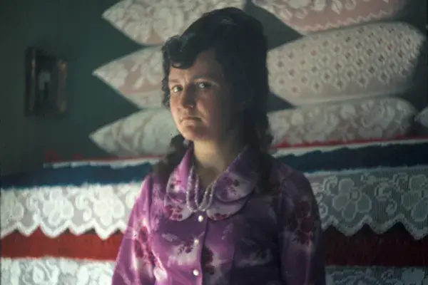 117.) Nő viseletben egy tisztaszobában, vetett ágy előtt. Mezőség, 1980-as évek